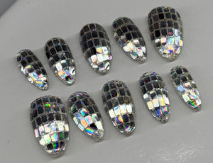 Ten oval-shaped clear acrylic press-on nails with a silver and black mosaic disco ball pattern, arranged on a light gray display stand.
