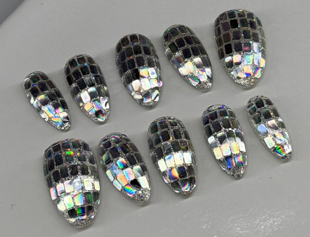 Ten oval-shaped clear acrylic press-on nails with a silver and black mosaic disco ball pattern, arranged on a light gray display stand.